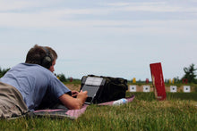 Load image into Gallery viewer, A person seated on grass is using a PlotMarker device to mark shot location; targets in the distance, which are part of a shooting range.
