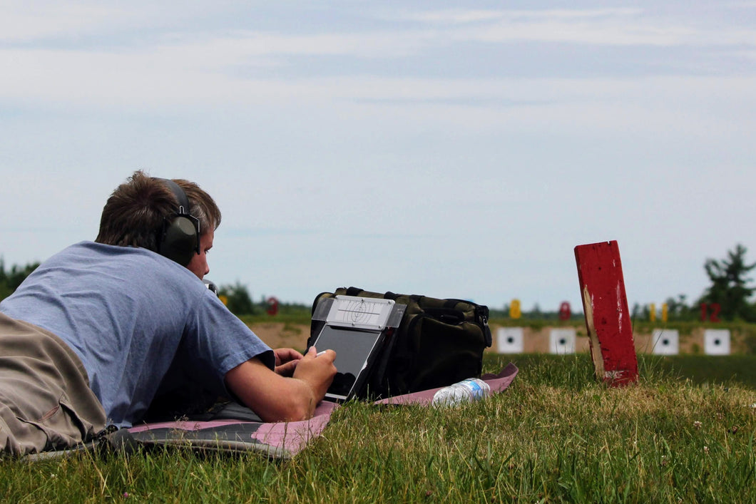 A person seated on grass is using a PlotMarker device to mark shot location; targets in the distance, which are part of a shooting range.